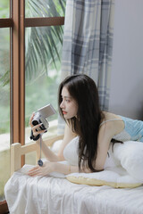 Young woman in light blue top lying on a bed, capturing memories with a camera by the window.
