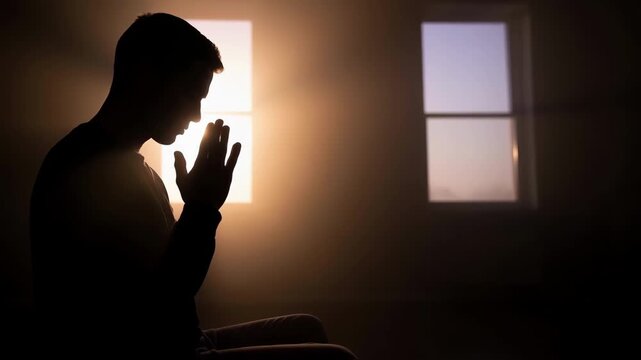 A silhouette of a man praying with hands together in a dark room against a bright window, representing the concept of faith, hope, spirituality, worship and mental health