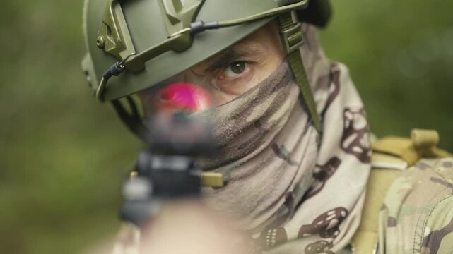 Camouflaged and masked army soldier with AR15 carbine. Rifle with a collimator aiming at the camera. Selective focus