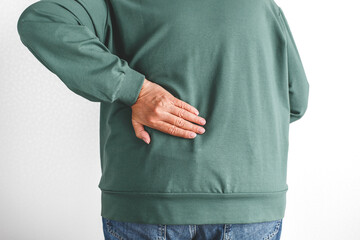 Close up of woman touching lower back with hand suffering from pain standing in casual green sweatshirt and jeans against light background