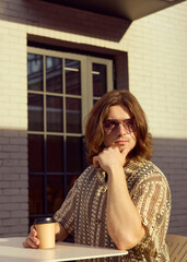 A thoughtful man in a stylish shirt and glasses drinks coffee from a paper cup on the street
