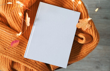 Top view of white book on wooden decorative table with fairy lights , leaves and yellow sweater
