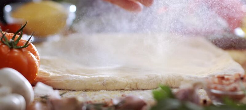 Close-up of a professional pizzaiolo's hands sprinkling white flour over freshly kneaded pizza dough on a kitchen counter surrounded by ingredients like tomatoes, mushrooms, and cheese