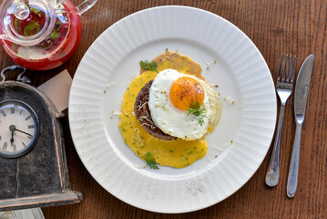 steak with sauce topped with fried egg on a plate on a table with tea and a retro clock