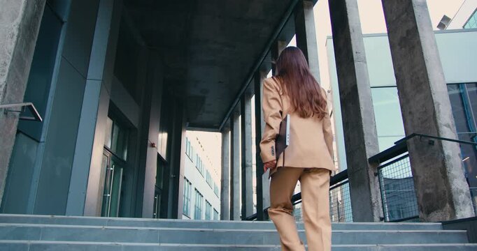 Rear view of confident businesswoman with long flowing hair, wearing tailored formal suit, walking up outdoor stairs while holding laptop in one hand, approaching modern office building
