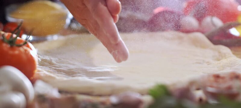 Close-up of a professional male baker kneading fresh dough on a wooden board, surrounded by ingredients like tomatoes and mushrooms, while sprinkling flour to prepare an authentic italian pizza