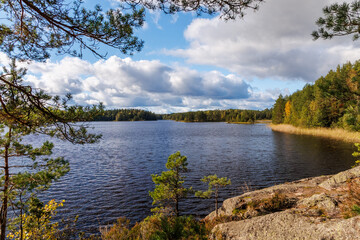 Scenic view of a calm lake surrounded by pine forest and rocky shore under a partly cloudy autumn sky, peaceful Scandinavian nature landscape ideal for travel, hiking, and outdoor exploration