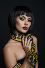 woman poses confidently with a large snake wrapped around her neck. Her dramatic makeup features bold colors, contrasting with the dark background, creating a striking visual