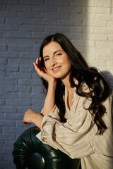 woman with long dark hair is sitting against a white brick wall, smiling gently while resting her chin on her hand in a warm, inviting atmosphere