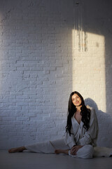 woman sits gracefully on the floor, stretching her legs in a bright studio. soft light from the window creates a warm atmosphere, highlighting the white walls and her relaxed pose