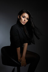 woman sits on a stool with long black hair flowing, dressed in a stylish black outfit, captured in dramatic lighting during a creative photo session