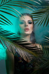 woman stands in a tropical environment, partially obscured by palm leaves and green smoke. Soft light enhances the vibrant colors and creates a mysterious atmosphere