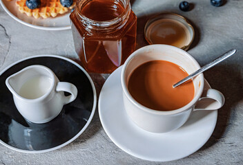 Coffee with milk in a cup and a jar of honey nearby, and next to it a jar of honey and blueberries