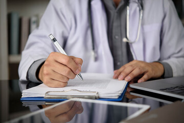 Doctor hand writing on medical clipboard, filling patient form at clinic desk. Physician in white coat with stethoscope writing prescription, working with laptop. Healthcare, medicine, 