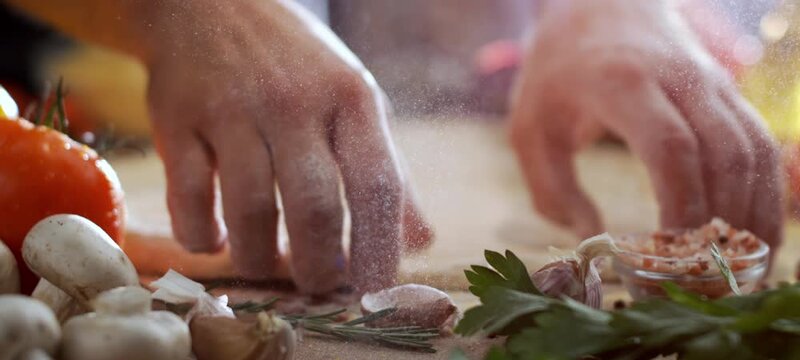 Vertical video of male chef's hands kneading and stretching fresh pizza dough on a wooden board covered in flour, with tomatoes, mushrooms, garlic, and herbs creating an authentic culinary scene