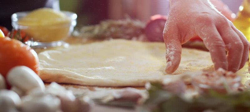 Vertical video of close-up of a baker's hands kneading and stretching fresh pizza dough on a floured wooden board, surrounded by fresh ingredients like tomatoes, mushrooms, and herbs in a kitchen