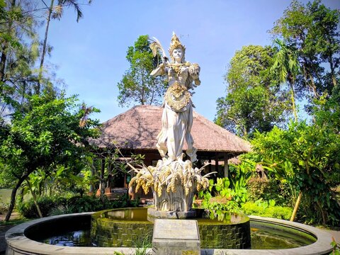  Mengwi, Badung, Bali, Indonesia, August 20, 2025: Balinese Dewi Sri statue, the goddess of rice and fertility, in a lush tropical garden setting with a traditional thatched roof pavilion. Culture, r