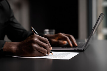 Close up of hands writing notes on printed business document while using laptop, showing focus and productivity in modern office environment