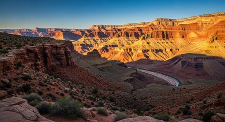 Scenic view of the Grand Canyon at sunset, showcasing the layered rock formations, Colorado River, and desert vegetation under a clear blue sky.