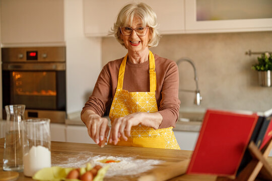 Portrait of happy elderly woman in apron breaking eggs into flour while creating cookie recipe blog post at home
