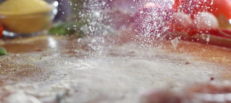 Vertical video of slow motion shot of white flour being thrown onto a rustic wooden tabletop, creating a dusty cloud with fresh cooking ingredients like tomatoes and onions arranged in the background