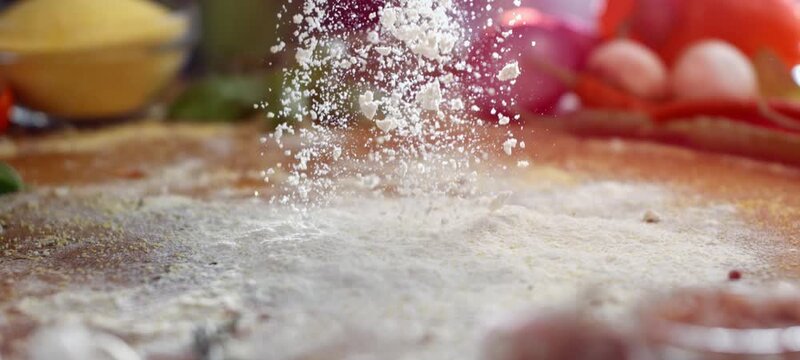 Close-up of a professional chef sprinkling white flour on a wooden kitchen counter, preparing to knead dough for pizza or bread with fresh ingredients in the background, showing the cooking process