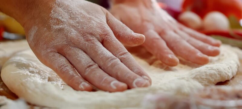 Close up view of a baker's hands kneading and stretching fresh pizza dough on a wooden board, preparing a traditional italian meal in a restaurant kitchen with ingredients in the background