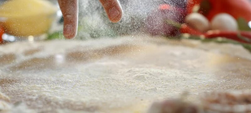 Vertical video of slow motion of white flour falling and spreading across a wooden kitchen counter, with fresh vegetables like tomatoes, garlic, and onions arranged in the softly blurred background