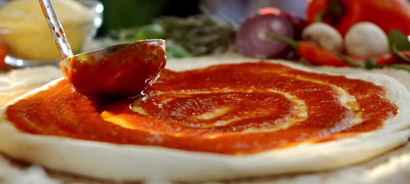 Vertical video of professional chef using a ladle to spread rich tomato sauce in a circular motion on fresh pizza dough, with fresh ingredients like mushrooms, onions, and peppers in the background