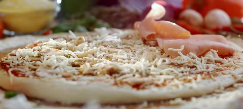 Delicate slices of cooked ham slowly falling onto a raw pizza base topped with tomato sauce and grated mozzarella cheese, showing the art of homemade italian food preparation in a kitchen