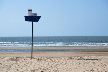 Wooden model of boat on pole on sandy beach with real big cargo ship in background, sunny day