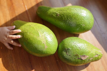 A trio of fresh, ripe avocados on a wooden surface, a child's hand reaching out to one of them