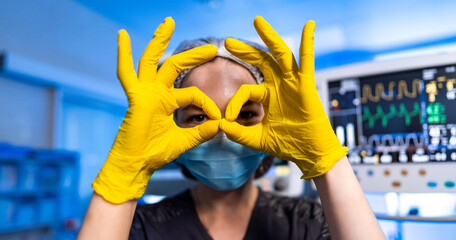 Nurse in yellow gloves near monitors. Nurse in black uniform and mask making funny gesture with yellow gloves near patient monitoring equipment in surgery room.