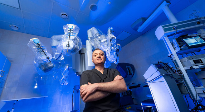 Medical specialist near robotic arms in surgical room. Healthcare worker in uniform with robotic arms prepared for surgery in modern hospital. - Powered by Adobe