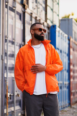 Black man in sunglasses and windbreaker jacket standing near cargo containers in port  © Dmytro Hai