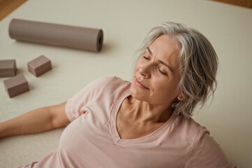 Elderly woman resting on yoga mat with eyes closed and relaxed expression after completing a home workout session with yoga equipment in background.