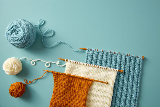 Flat lay of colorful knitting samples in blue, white, and orange yarn on wooden needles. Handmade craft and creative textile work.