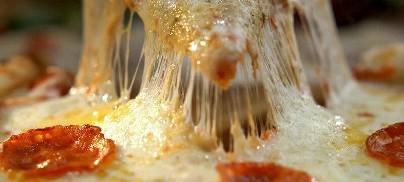 Macro shot of a person taking a slice of hot pepperoni pizza with delicious melted mozzarella cheese stretching in an appetizing and stringy way, showing the greasy and tasty ingredients