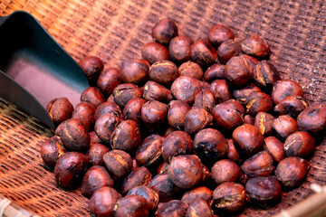 Close-up of roasted chestnuts with cracked, glossy shells in a wicker basket. A metal scoop in the corner hints at serving, with warm lighting creating a cozy, autumn street food vibe.