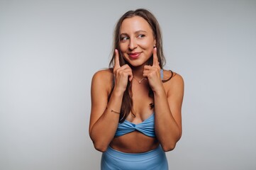 Fototapeta premium Smiling woman playfully posing with her fingers on her cheeks in a light blue outfit in a bright studio setting