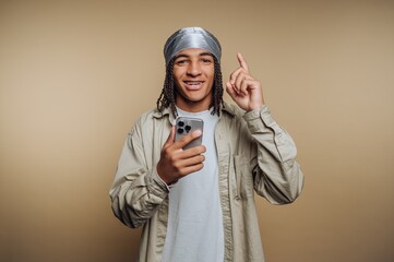 Young man in beige shirt and silver headscarf smiles while holding a smartphone indoors