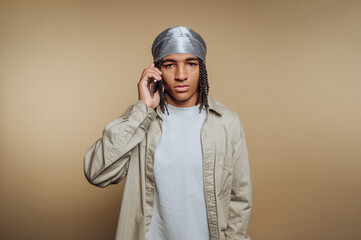 Young man wearing silver head wrap talks on mobile phone against neutral background in casual outfit