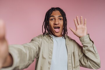 Young man smiling and waving happily against a pink background in a casual setting with bright lighting