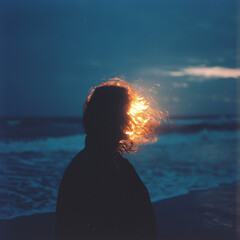 Silhouette of a person by the ocean at dusk with sunlight shining through their hair.