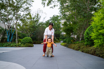 Asian grandmother teaching her grandson to walk outdoors during summer. A family with children, showcasing the lifestyle between grandparents and grandchildren.