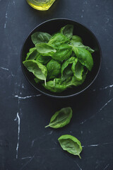 Bowl with fresh green basil leaves on a black marble background, vertical shot, top view