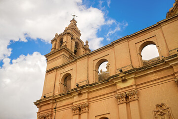 Close-up of an old church tower with bell, weather vane, carved stone details, arched windows, and classical design under a blue cloudy sky 