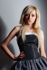 Blonde beautiful young woman wearing a chic black dress holds a confident pose and bites her lip against a professional studio background, highlighting elegance and modern style.