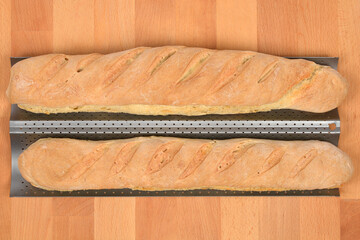 An overhead shot features two freshly baked, golden-brown baguette loaves resting in a perforated metal baking pan against a light wooden kitchen countertop background