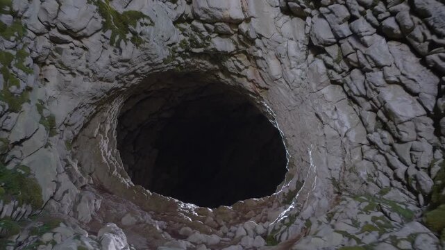 Cave entrance opens into underground darkness. Large round hole carved in rock wall. Stone surface shows natural formation. Shadows fall inside deep cave space with rugged rock and stone textures